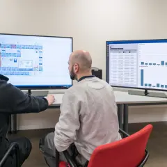 Dwóch mężczyzn analizuje dane na dwóch monitorach: jeden z planem, drugi z wykresem słupkowym.