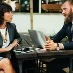 Kobieta i mężczyzna w garniturach rozmawiają przy stoliku z laptopem i tabletem. Może to być rozmowa o tym, dlaczego szukam nowej pracy.