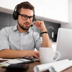 Mężczyzna w słuchawkach z mikrofonem pracuje zdalnie w USA z polski, siedząc przy laptopie w kuchni.