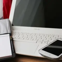 Biały laptop, notatnik z długopisem i smartfon. Czas pomyśleć o podatkach programisty i uporządkować finanse.