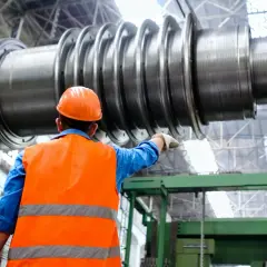 Pracownik w pomarańczowej kamizelce i kasku wskazuje na ogromny element turbiny. Dobry technolog produkcji może liczyć na wysokie zarobki.