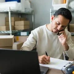 Przedsiębiorca rozmawia przez telefon, notując dane. Zastanawia się nad wyborem formy opodatkowania, np. ryczałt, i czy przekroczy próg podatkowy.