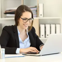 Uśmiechnięta kobieta w okularach pracuje na laptopie, planując swoje samozatrudnienie w Polsce.