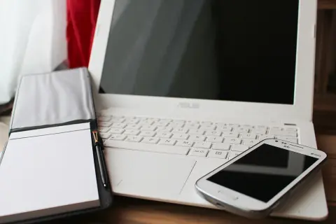 Biały laptop, notatnik z długopisem i smartfon. Czas pomyśleć o podatkach programisty i uporządkować finanse.