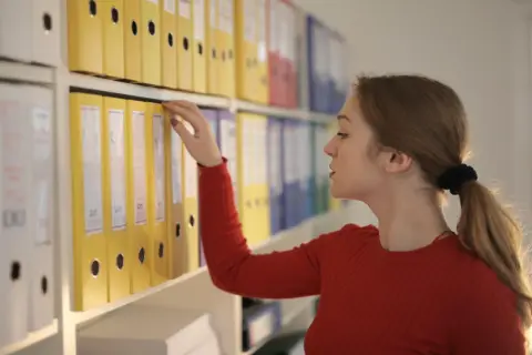 Kobieta w czerwonym swetrze sięga po żółty segregator na półce w biurze. Dokumenty uporządkowane, to klucz do efektywnej pracy w lean office.