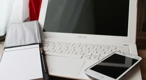 Biały laptop, notatnik z długopisem i smartfon. Czas pomyśleć o podatkach programisty i uporządkować finanse.