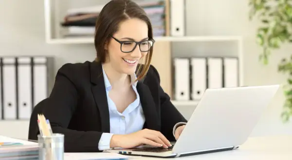 Uśmiechnięta kobieta w okularach pracuje na laptopie, planując swoje samozatrudnienie w Polsce.