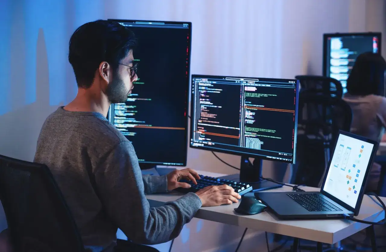 Młody b2b programista w okularach pracuje nocą przy wielu monitorach, pisząc kod i analizując schematy.