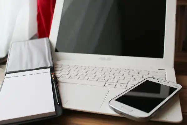 Biały laptop, notatnik z długopisem i smartfon. Czas pomyśleć o podatkach programisty i uporządkować finanse.