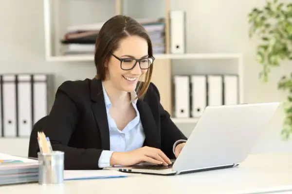 Uśmiechnięta kobieta w okularach pracuje na laptopie, planując swoje samozatrudnienie w Polsce.